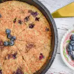 keto blueberry toasted coconut skillet cake in cast iron skillet with dish of blueberries and cake server on the right