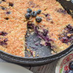 keto blueberry toasted coconut skillet cake in cast iron skillet with one slice missing and dish of blueberries