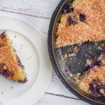 keto blueberry toasted coconut skillet cake in cast iron skillet with one slice missing and slice on white plate on the left